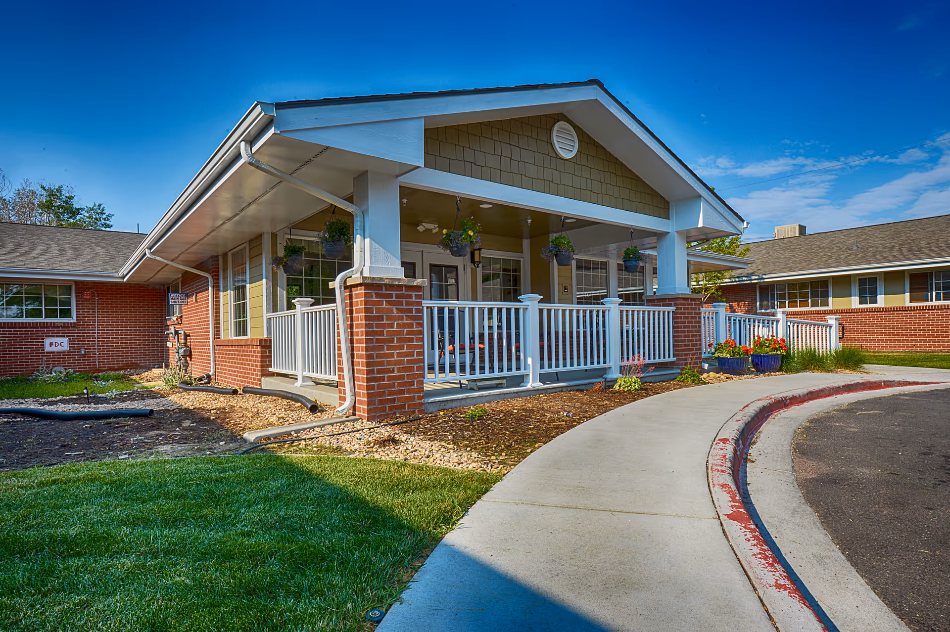 Exterior view of a single-story brick building with a covered porch featuring white railings and hanging flower pots. The building is surrounded by a curved sidewalk, green grass, and flower beds under a clear blue sky.