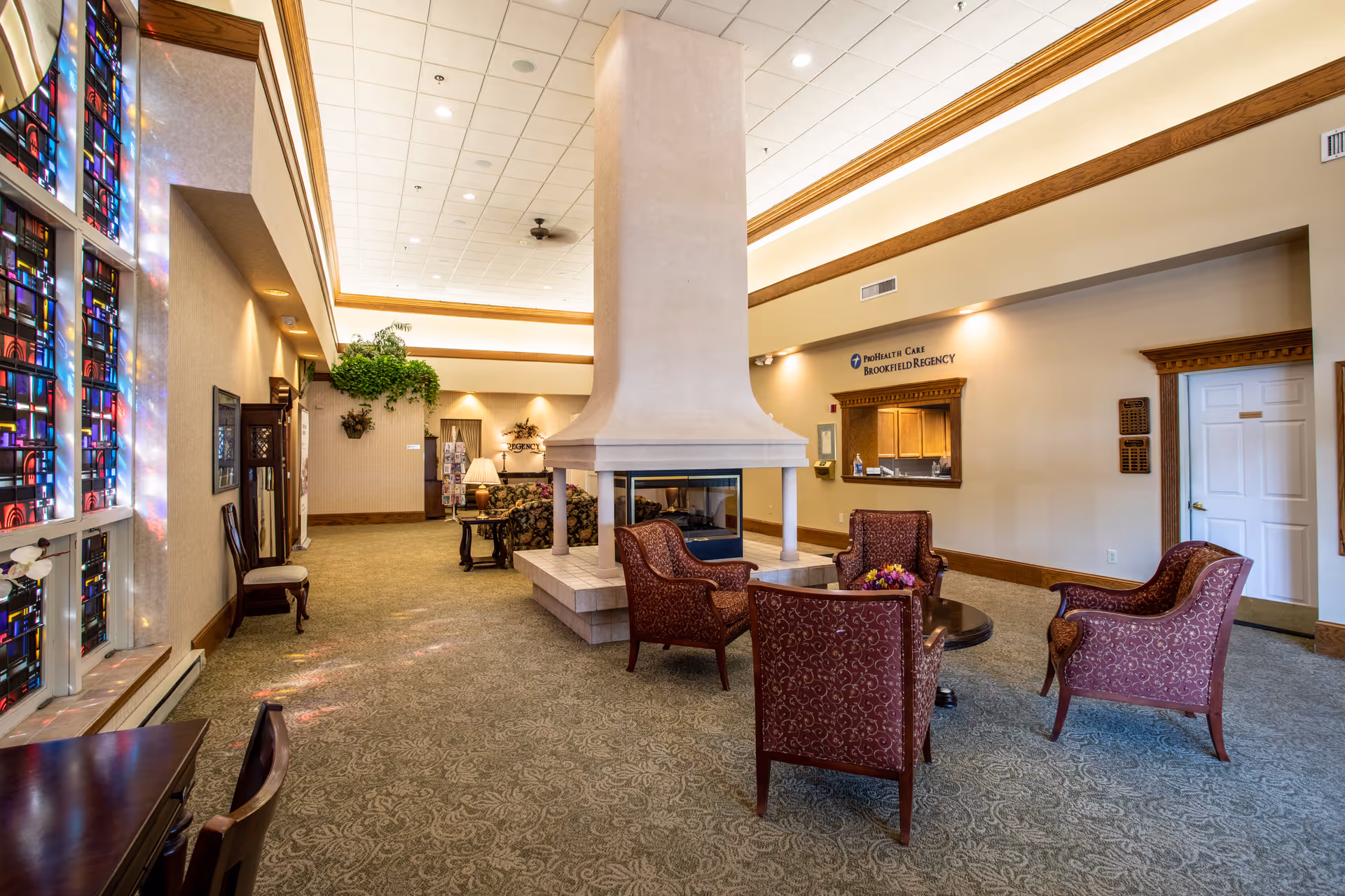 A spacious senior living community lounge area with a central double-sided fireplace surrounded by four upholstered chairs and a small round table with a flower arrangement. The room features patterned carpet, a high ceiling with recessed lighting, stained glass windows on the left, and a reception window on the right wall with the sign 'ProHealth Care Brookfield Regency'. There are decorative plants and lamps in the background, creating a warm and inviting atmosphere.