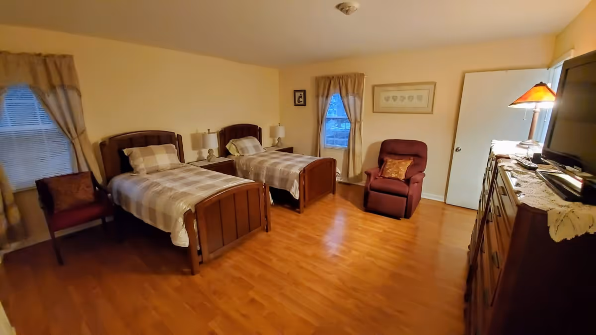 A bedroom with two single beds featuring wooden headboards and plaid bedding. Between the beds is a nightstand with two lamps. There are two windows with beige curtains, a burgundy armchair with a decorative pillow, a wooden dresser with a lace runner, a lamp, and a television on top. The room has wooden flooring and light-colored walls.