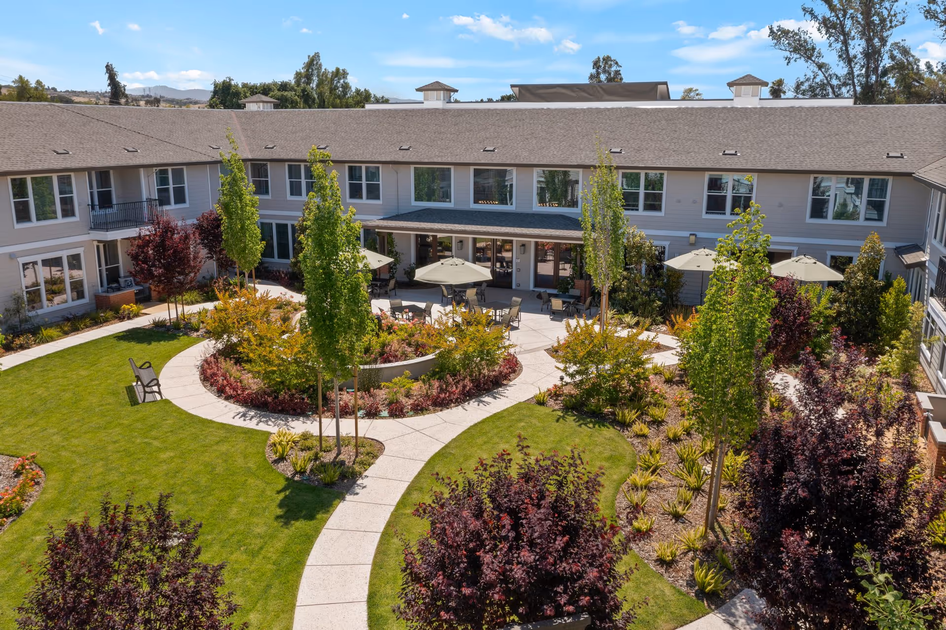 A landscaped courtyard at Oakmont of Silver Creek featuring green lawns, walking paths, various trees and shrubs, outdoor seating with tables and umbrellas, and a two-story building surrounding the courtyard under a blue sky.