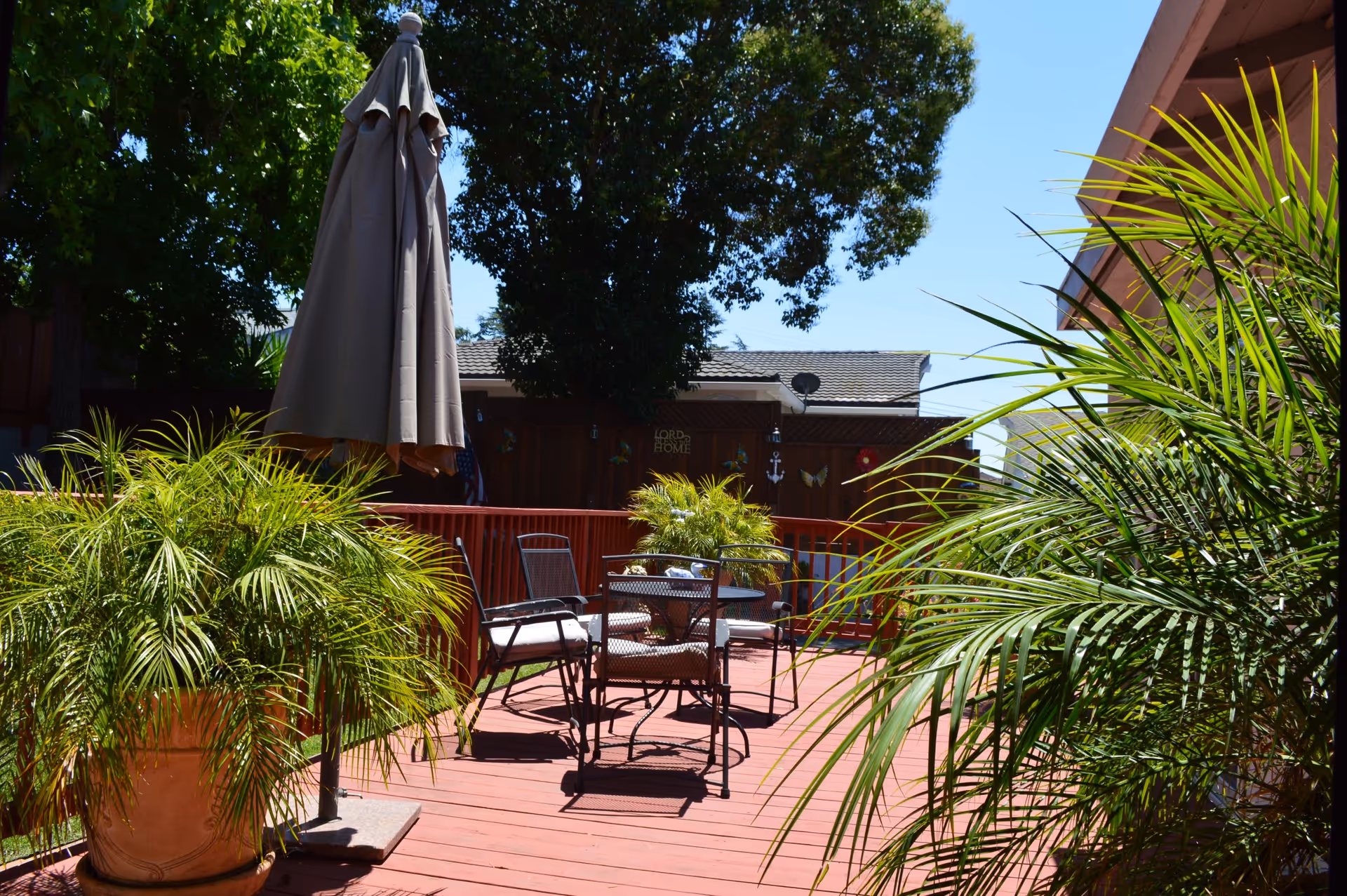 Outdoor patio area with a red wooden deck, metal table and chairs with cushions, a closed beige umbrella, and potted green plants. Trees and a building are visible in the background under a clear blue sky.
