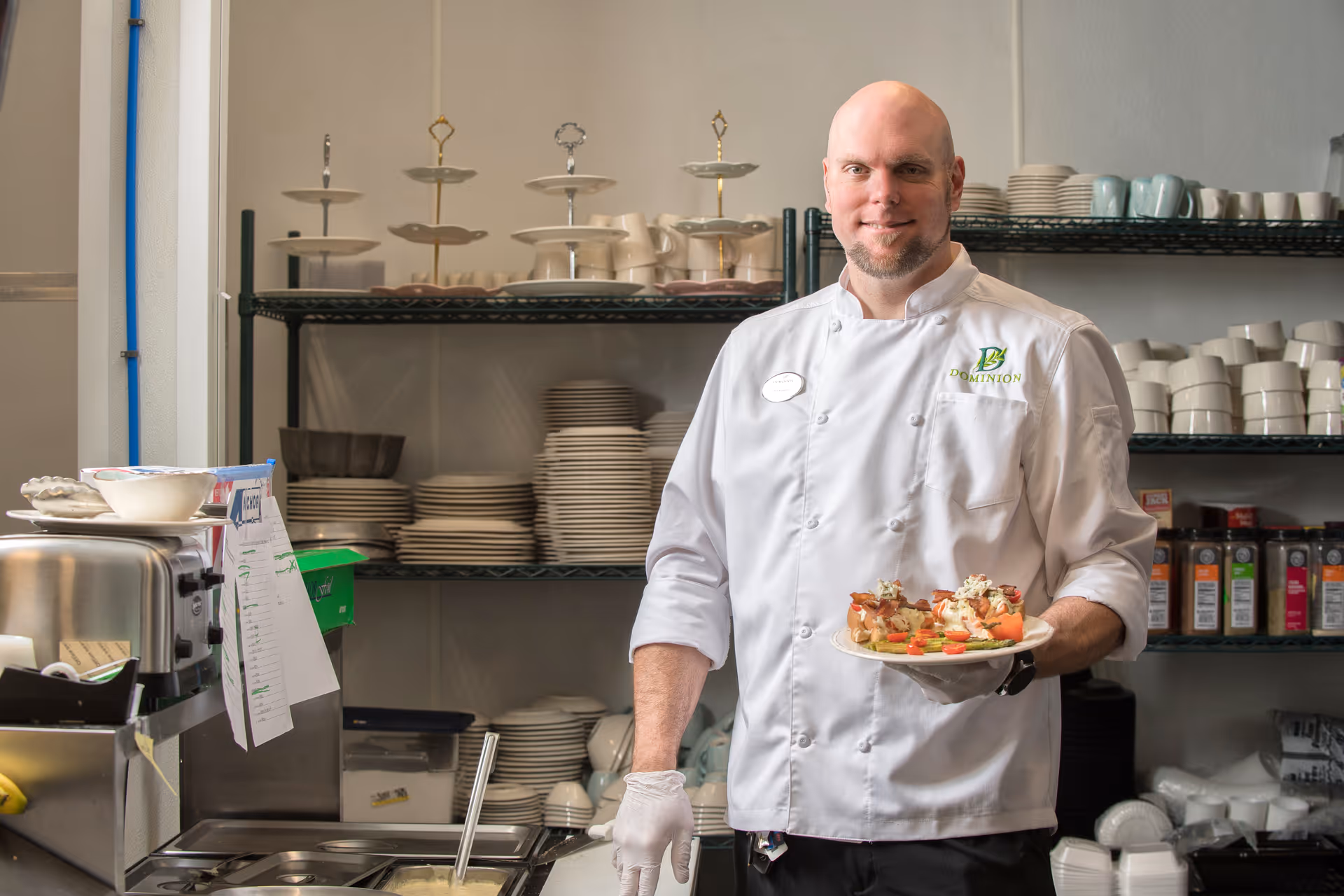 A chef wearing a white uniform with the Dominion logo stands in a kitchen holding a plate of prepared food. Behind him are shelves stocked with plates, bowls, cups, and various kitchen items.