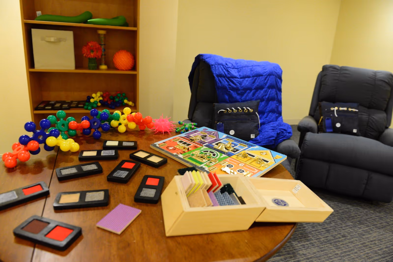 A therapy or activity room with a wooden table covered in various sensory and tactile items including colorful plastic molecular models, texture sample cards, and a board with different types of locks. Two dark recliner chairs are in the background, one draped with a blue blanket and a black sensory bag. A wooden shelf with more sensory items and a small vase with a red flower is visible against the wall.