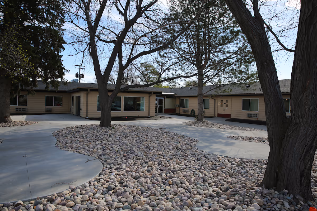 Outdoor courtyard area of Monte Vista Hills Health Care Center featuring leafless trees surrounded by a bed of small rocks and concrete walkways, with the building visible in the background under a clear sky.