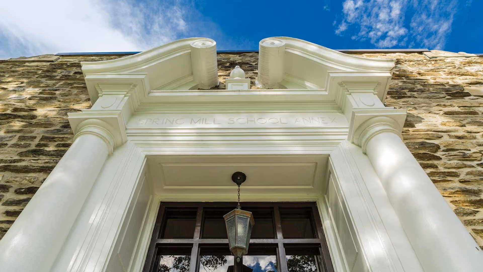 Low-angle view of a stone building entrance with white columns, a decorative pediment engraved "SPRING MILL SCHOOL ANNEX" and a hanging lantern.