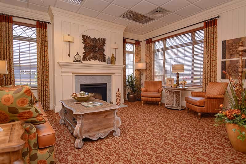 A cozy living room area with a patterned carpet, a wooden coffee table, a fireplace with decorative items on the mantel, two armchairs with a small round table between them, large windows with blinds and patterned curtains, and a sofa with floral cushions.