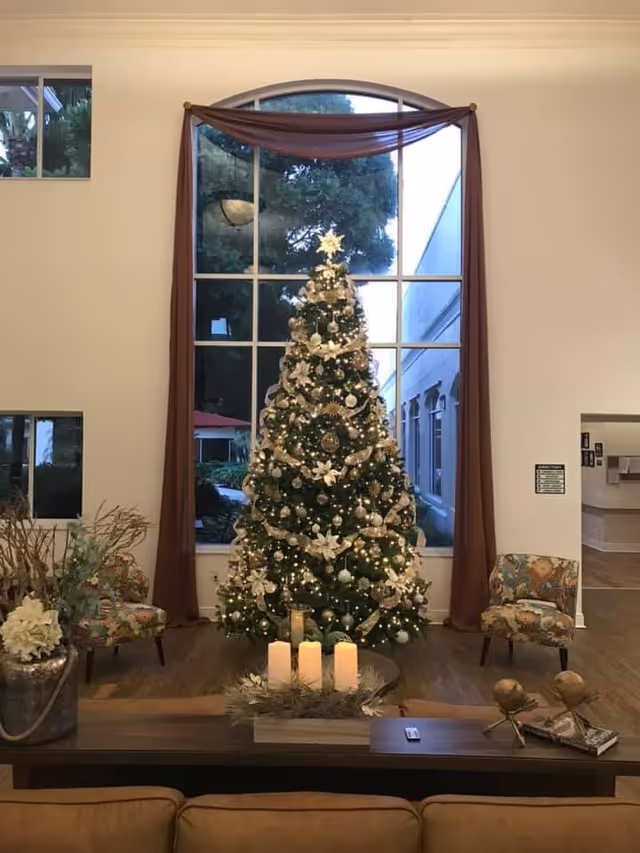 A decorated Christmas tree with white and gold ornaments and lights stands in front of a large arched window with brown curtains in a cozy living room. Two floral upholstered chairs are placed on either side of the tree. In the foreground, a wooden table holds a centerpiece with four lit candles and decorative items. The room has wooden flooring and light-colored walls.
