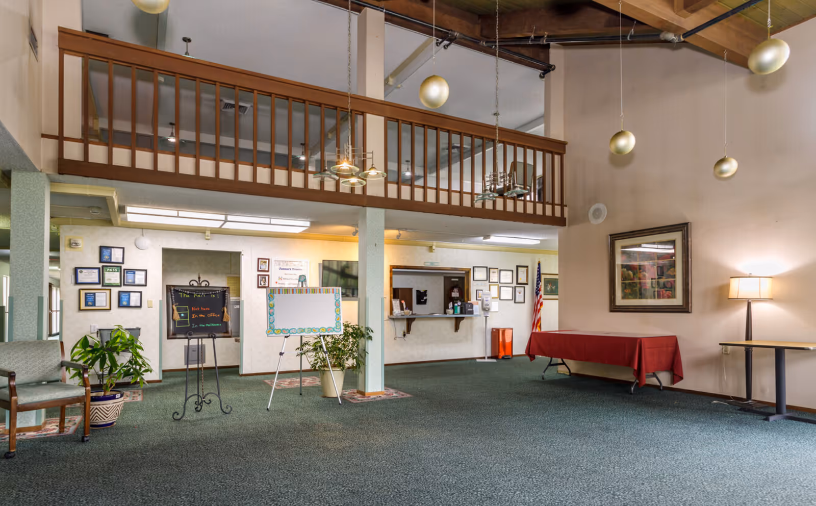 Spacious senior living facility lobby with high ceilings, balcony railing, seating, plants, bulletin boards, an American flag and a reception window.