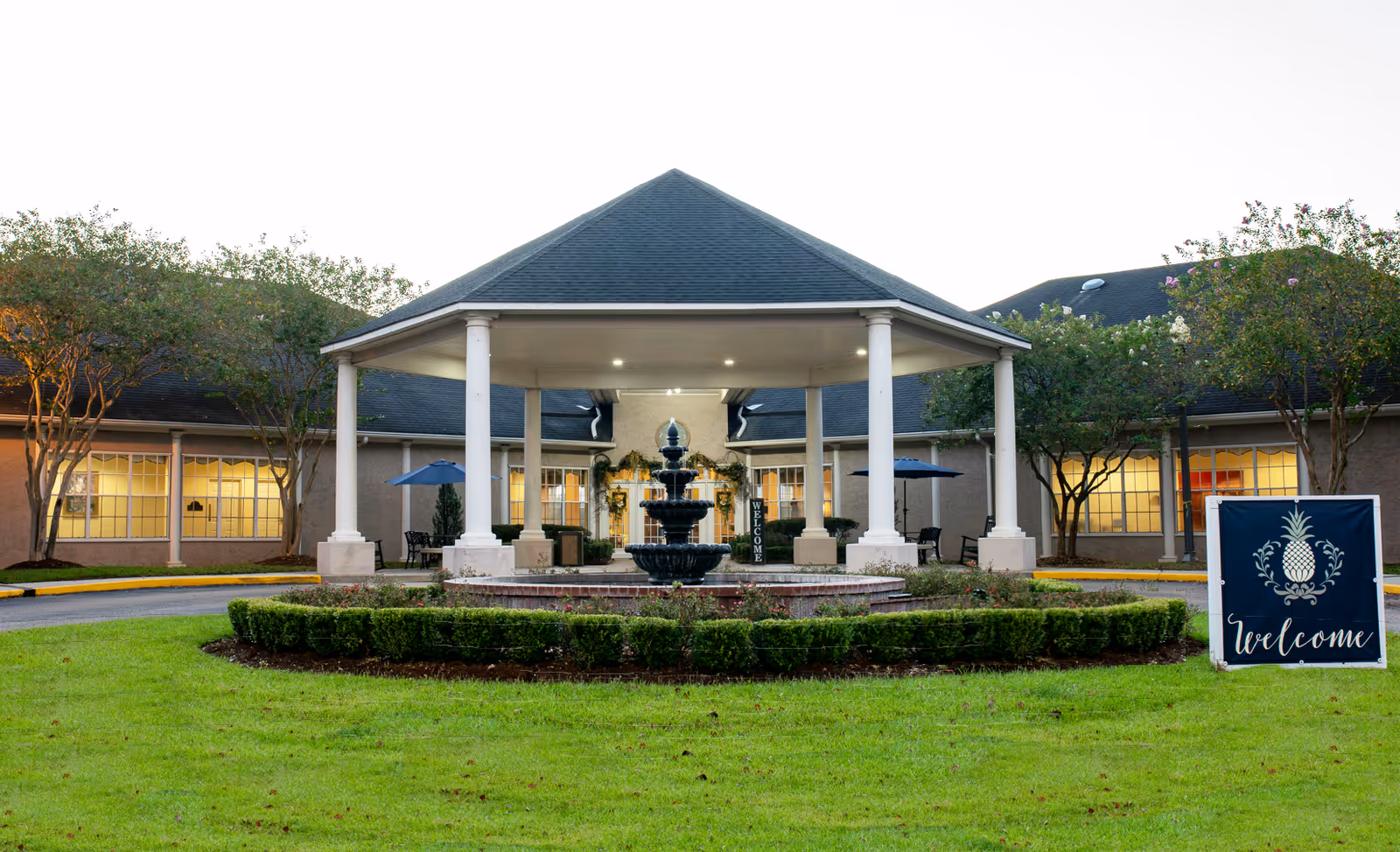 Exterior view of Lake Sherwood Village Retirement Community featuring a covered entrance with white columns, a central black tiered fountain, manicured bushes, green lawn, and a welcome sign with a pineapple design. The building has large windows and is surrounded by trees.