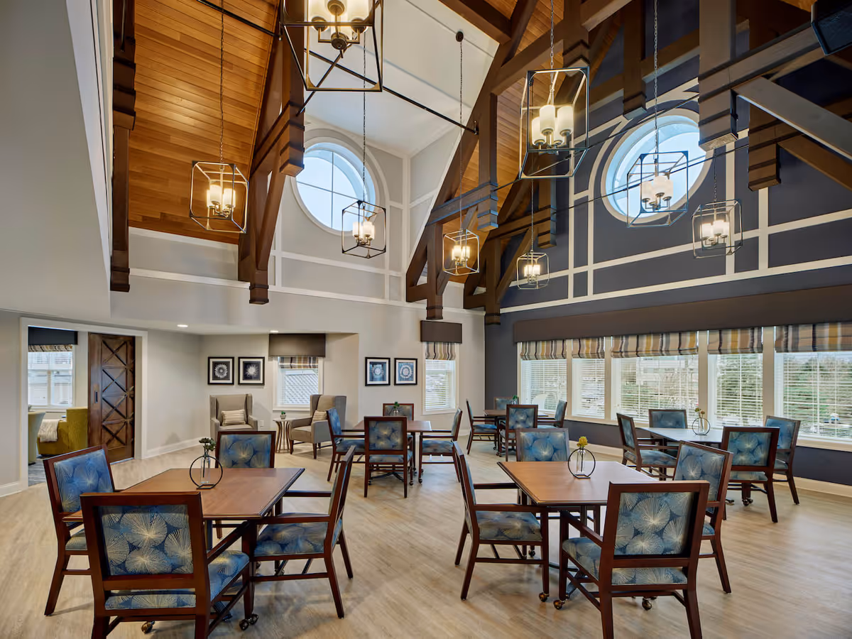 A spacious dining room with high vaulted wooden ceilings and large circular windows. The room features several wooden tables with blue patterned chairs arranged neatly. Modern pendant lights hang from the ceiling, and there are large windows with striped valances letting in natural light. In the corner, there are two armchairs with a small table between them and framed artwork on the walls.