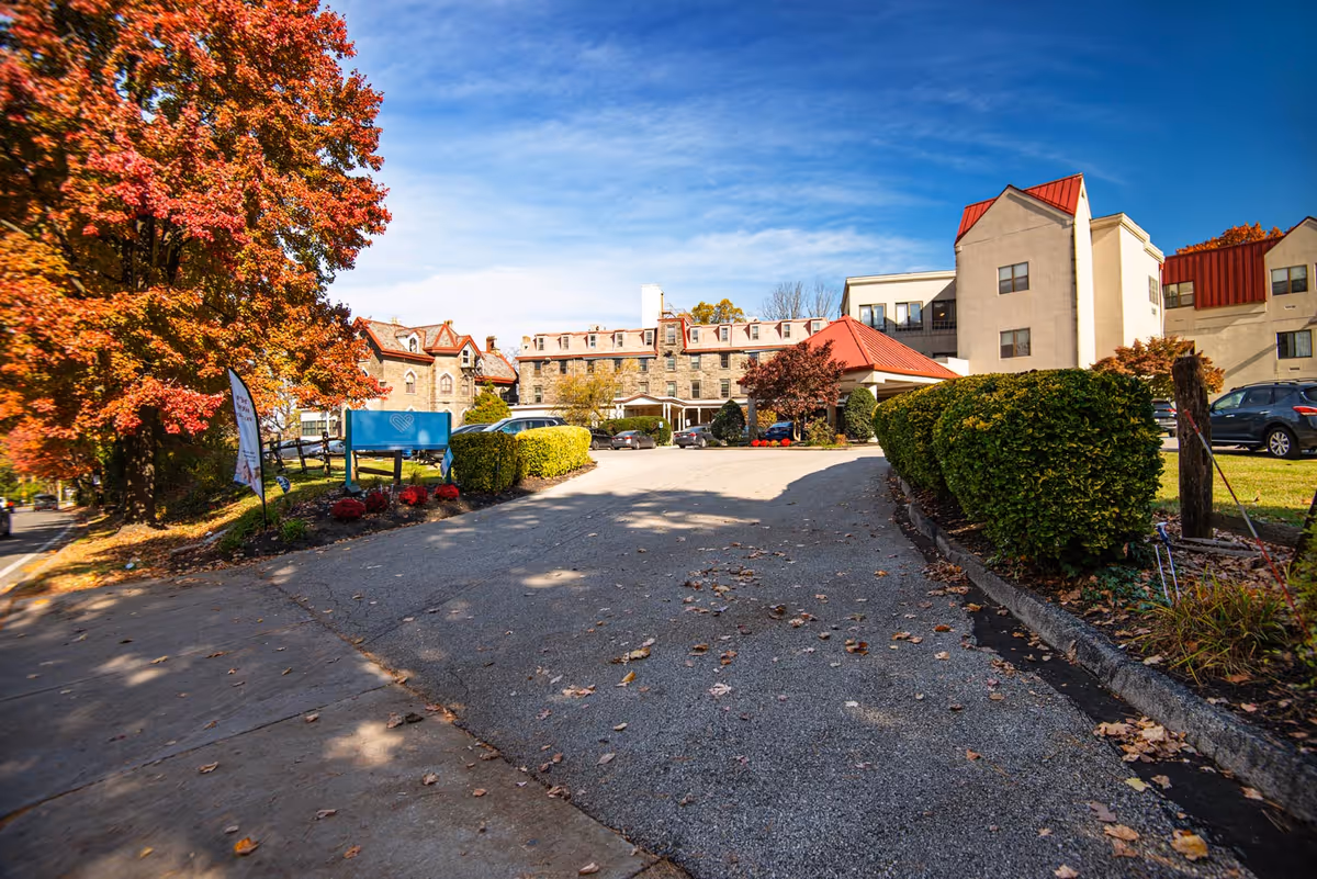 Exterior view of Fairview Nursing and Rehabilitation Center on a sunny day with a clear blue sky. The building features a mix of stone and beige walls with red roofs. There are several cars parked near the entrance, and the driveway is lined with trimmed bushes and autumn-colored trees.