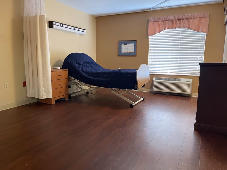 A senior care facility bedroom with a hospital-style adjustable bed covered with a dark blue blanket. The room has wooden flooring, a small wooden nightstand with drawers next to the bed, a window with closed blinds and a pink valance, and an air conditioning unit below the window. A privacy curtain is partially visible on the left side of the image.