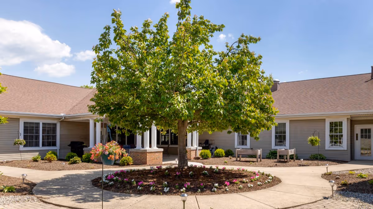 Courtyard entrance of Brookdale Harrisburg with a central tree surrounded by flower beds, walkways, benches and a single-story building.