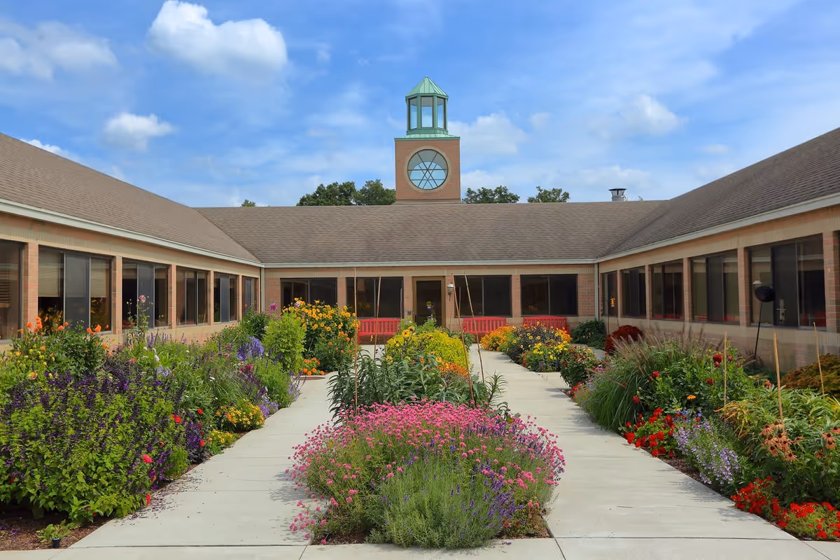 A courtyard garden at Chelsea Retirement Community with colorful flowers and plants lining a concrete walkway. The building surrounds the courtyard with large windows and a central tower featuring a decorative circular window and a green cupola under a partly cloudy blue sky.