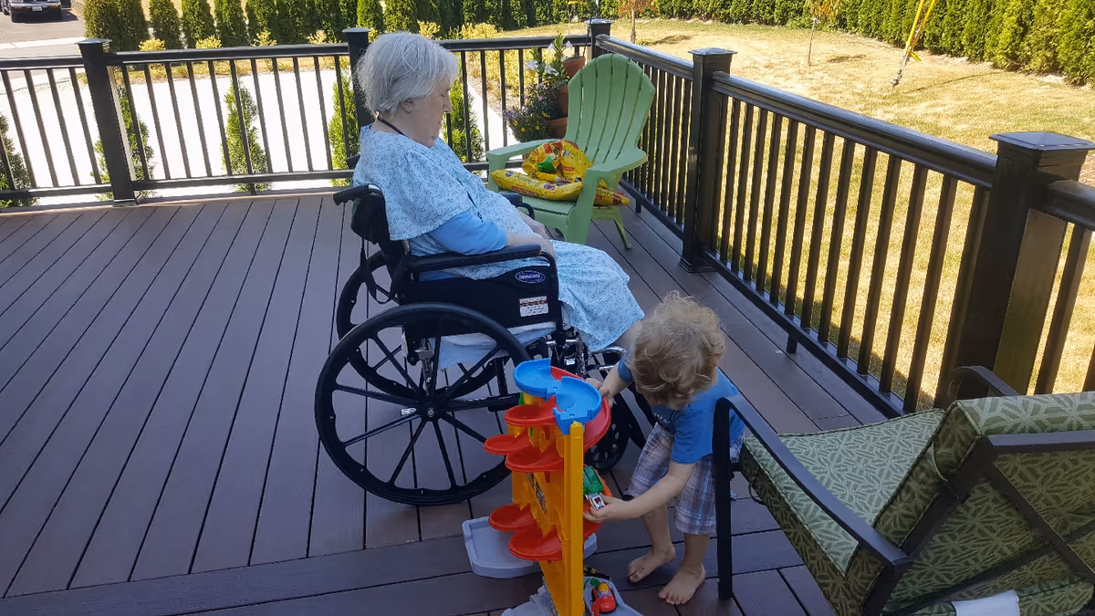 An elderly woman in a wheelchair on a wooden deck outside, accompanied by a young child playing with a colorful toy car ramp. The deck is surrounded by a black railing and has outdoor chairs, with a grassy area and trees visible in the background.