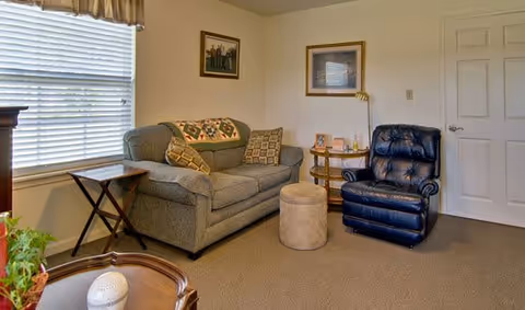 A cozy living room with a gray couch adorned with patterned pillows and a quilt draped over the back. Next to the couch is a small wooden side table and a round wooden table with a lamp, picture frames, and decorative items. A dark blue leather recliner chair is positioned near a closed white door. The room has beige walls, a large window with white blinds, and carpeted flooring.