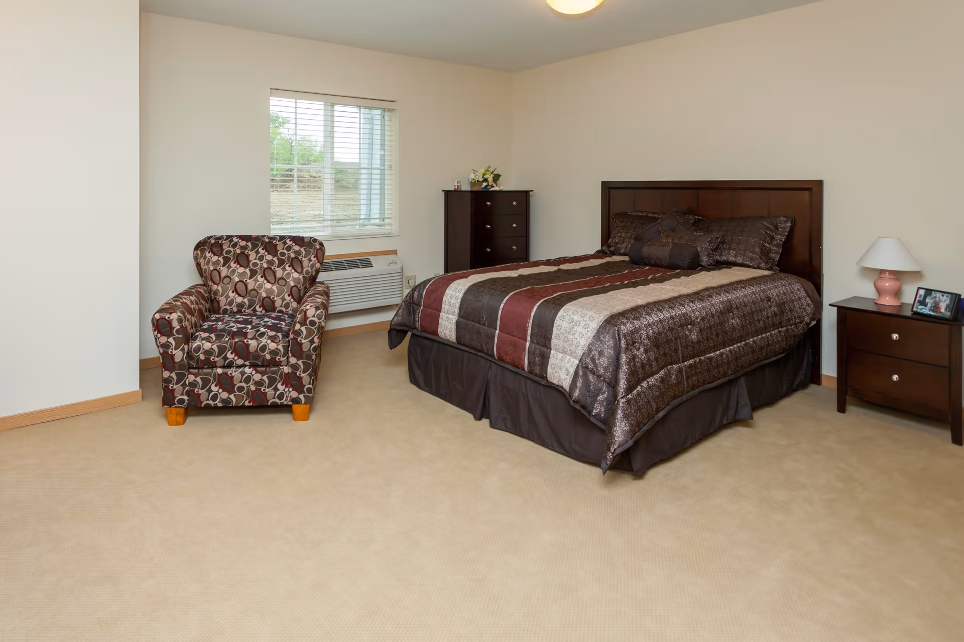 A bedroom with a large bed covered in a striped brown, beige, and maroon comforter, a patterned armchair, a dark wooden dresser, and a matching nightstand with a pink lamp and a framed photo. There is a window with blinds and a wall-mounted air conditioning unit beneath it.