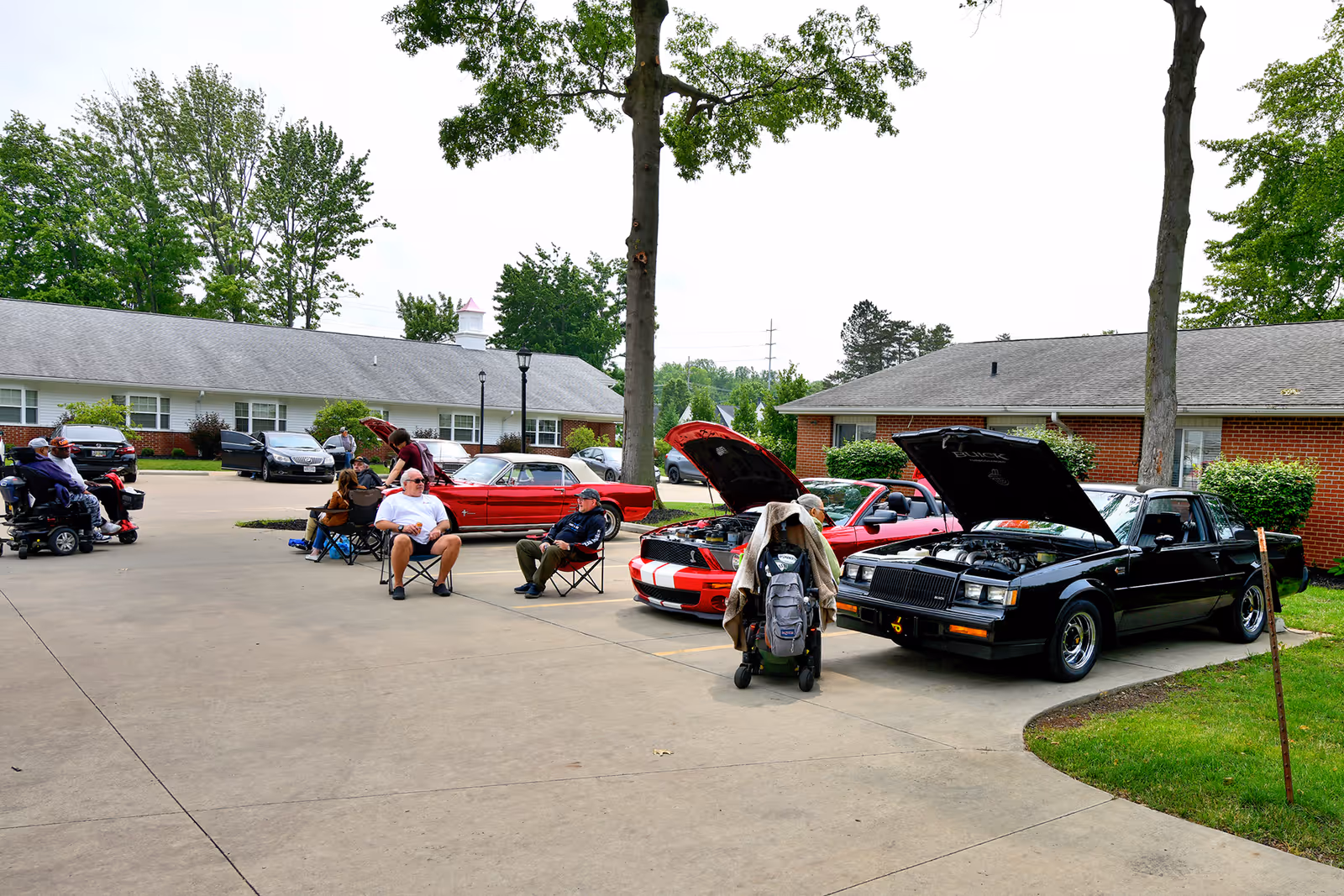 Residents sit and socialize in a courtyard while classic cars with open hoods are displayed outside a senior living facility.