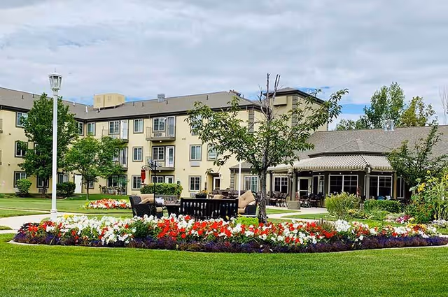 Outdoor garden area at Cedarwood at Sandy featuring a well-maintained lawn, colorful flower beds with red, white, and purple flowers, several trees, outdoor seating with cushioned chairs and tables, and a multi-story beige building in the background under a partly cloudy sky.