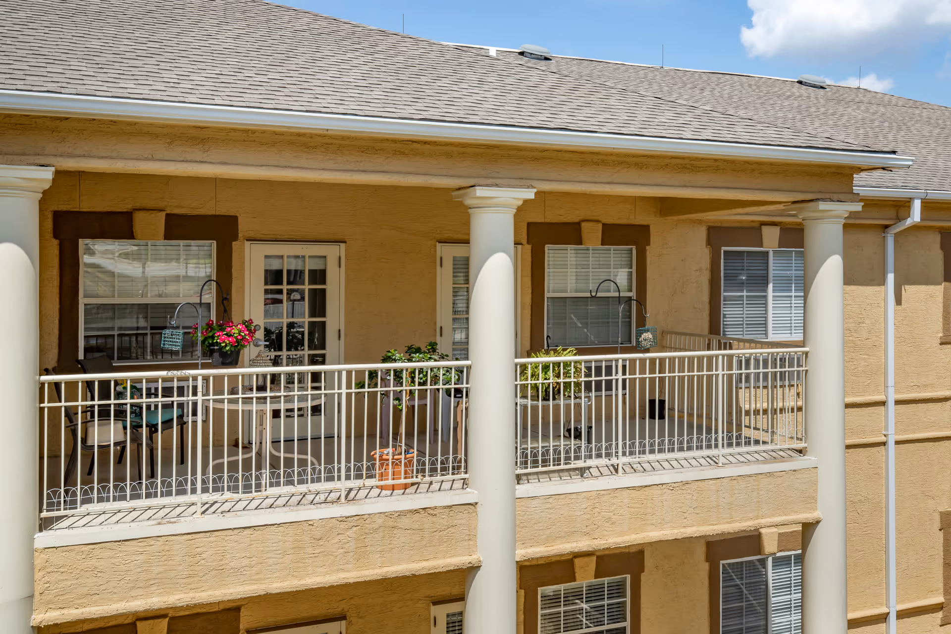 Exterior view of a balcony at Independence Hill Retirement Community with beige walls, white railing, two white columns, potted plants, and outdoor chairs. The balcony has two windows and a glass door leading inside.