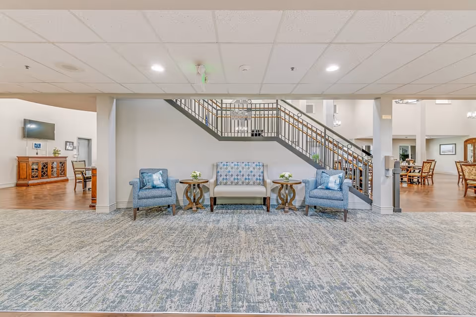 A spacious senior living facility common area with a patterned carpet floor and a staircase with metal railings. In front of the staircase, there is a seating arrangement consisting of a cushioned bench with a patterned backrest flanked by two blue armchairs, each with a small side table holding a flower arrangement. The area is well-lit with ceiling lights, and to the left and right, there are dining areas with wooden floors, tables, and chairs.