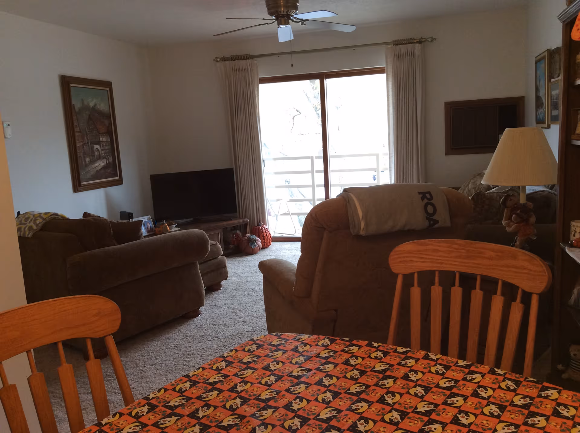 View of a cozy living room with brown upholstered chairs and a sofa, a TV on a wooden stand, a ceiling fan, and a sliding glass door leading to a balcony. In the foreground, there is a wooden dining table with chairs and a colorful tablecloth featuring a Halloween-themed pattern. The room is decorated with framed paintings and a table lamp.