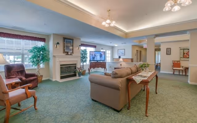 A spacious living room area in a senior living facility with green carpet, a beige sofa, wooden chairs, a fireplace, large windows with blinds and valances, potted plants, a TV on a wooden stand, and soft overhead lighting.