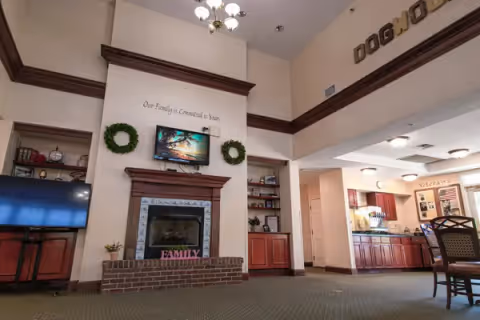 Common area of a senior living facility showing a fireplace with a mounted TV, built-in shelving and a kitchenette in the background.