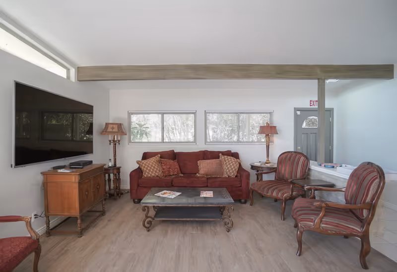 A cozy living room with a red sofa adorned with patterned cushions, two striped armchairs, a wooden coffee table with magazines, a wooden sideboard with a large flat-screen TV mounted above it, and two table lamps on either side of the sofa. The room has light-colored walls, wooden flooring, and two windows letting in natural light. A gray door with an exit sign is visible in the background.