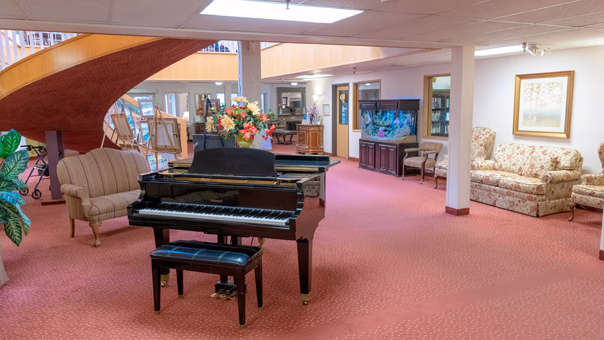 Spacious senior living common area with a grand piano, seating, an aquarium, and a curved staircase.