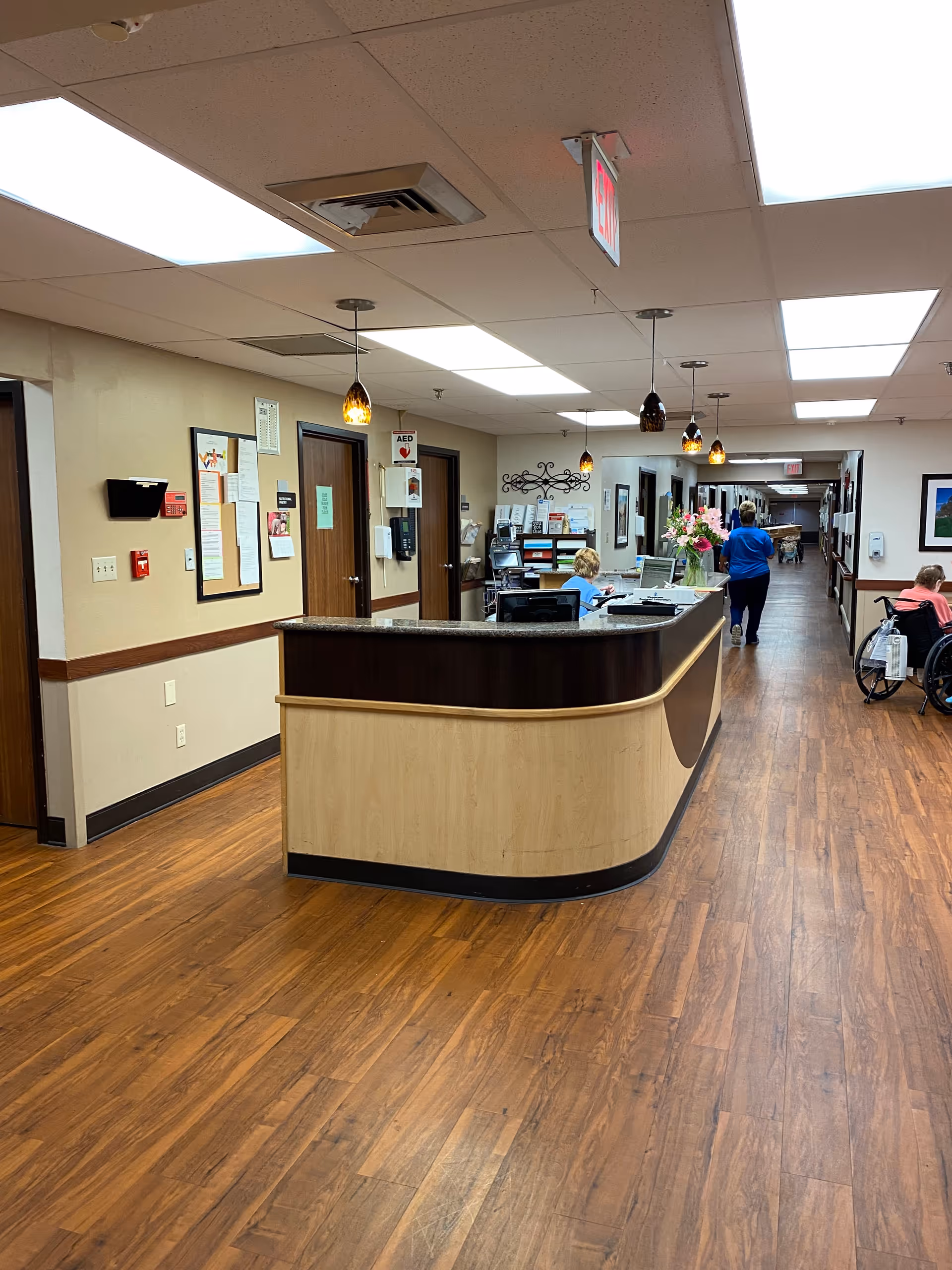 Interior view of a healthcare facility hallway with a wooden reception desk in the foreground. A staff member is seated behind the desk working on a computer, while another person in blue scrubs walks down the hallway. On the right side, a person in a wheelchair is visible. The hallway has wooden flooring, beige walls with a wooden trim, and multiple doors along the corridor. Ceiling lights and hanging pendant lights illuminate the area.