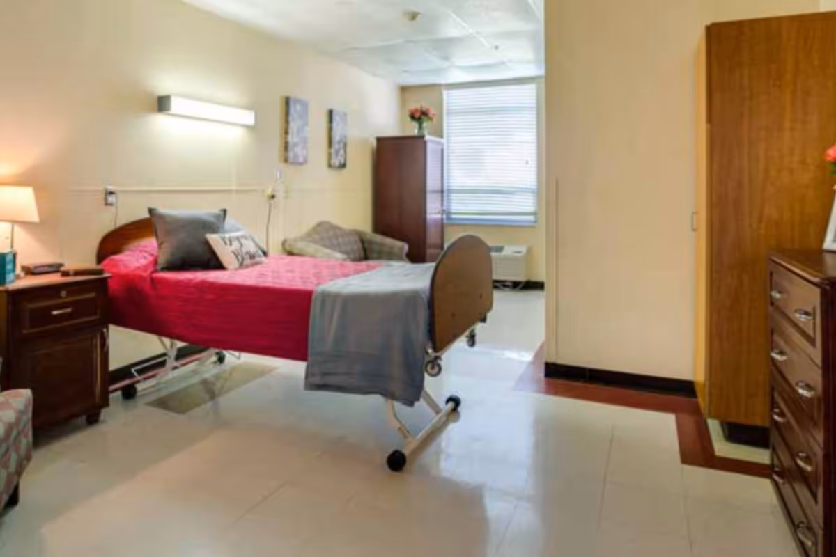 A senior living private room with a medical bed dressed in a red coverlet, bedside table, armchair, dresser and a window.