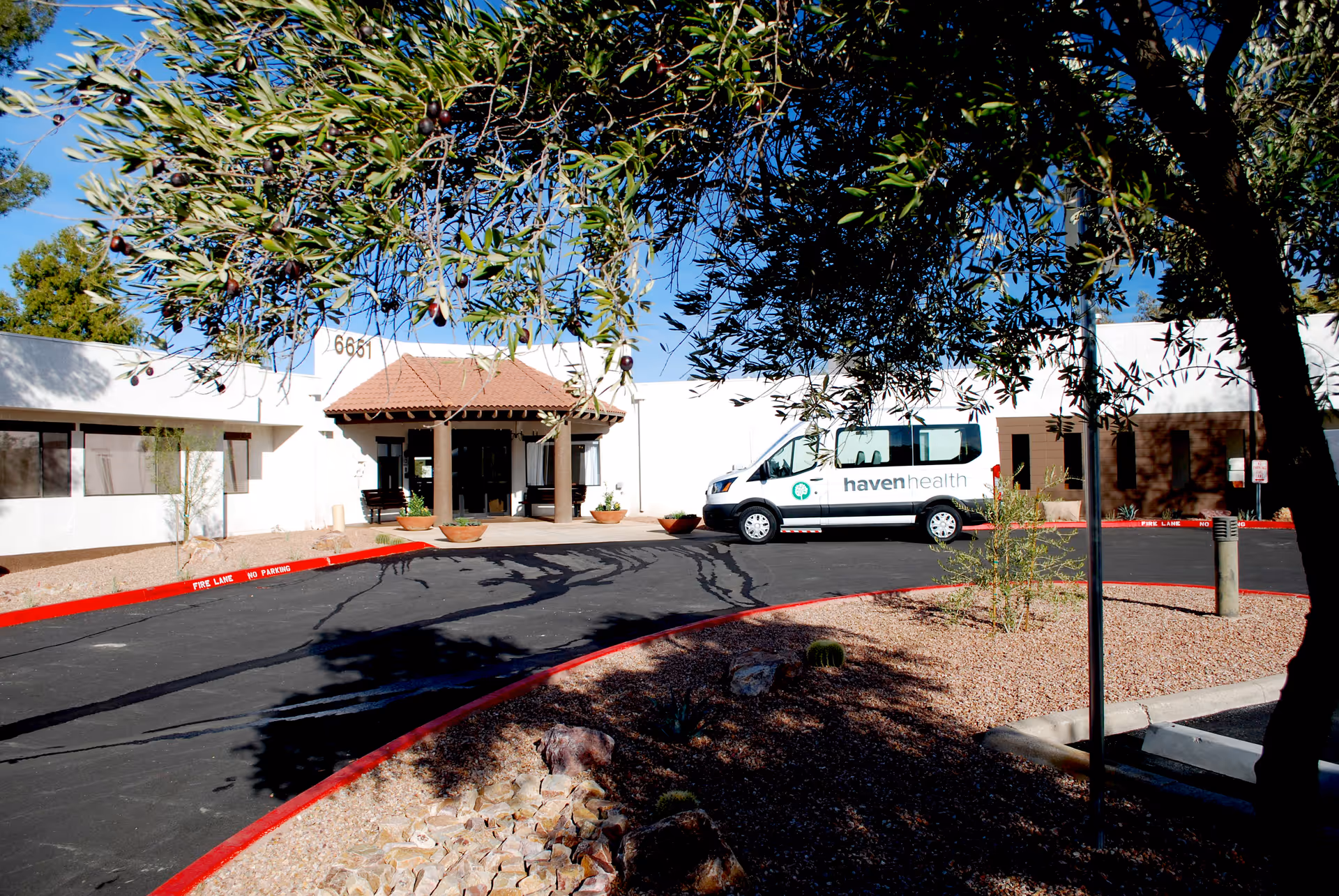Exterior view of Haven Health Saguaro Valley facility with a white building, a covered entrance with a tiled roof, and a white van parked in front displaying the Haven Health logo. The foreground includes desert landscaping with rocks, small plants, and a tree with green leaves and olives.