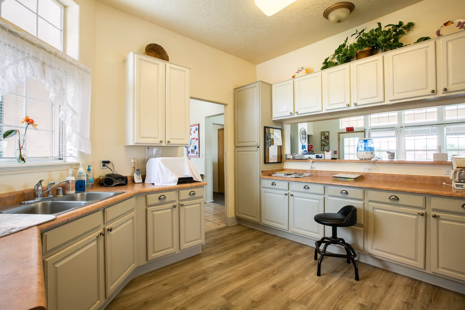 A bright kitchen area with cream-colored cabinets and wood-toned countertops. There is a double sink under a window with lace curtains, a black stool in front of the counter, and various kitchen items including a coffee maker, a covered appliance, and a water dispenser. The floor is wood-patterned, and there are plants and decorative items on top of the cabinets.