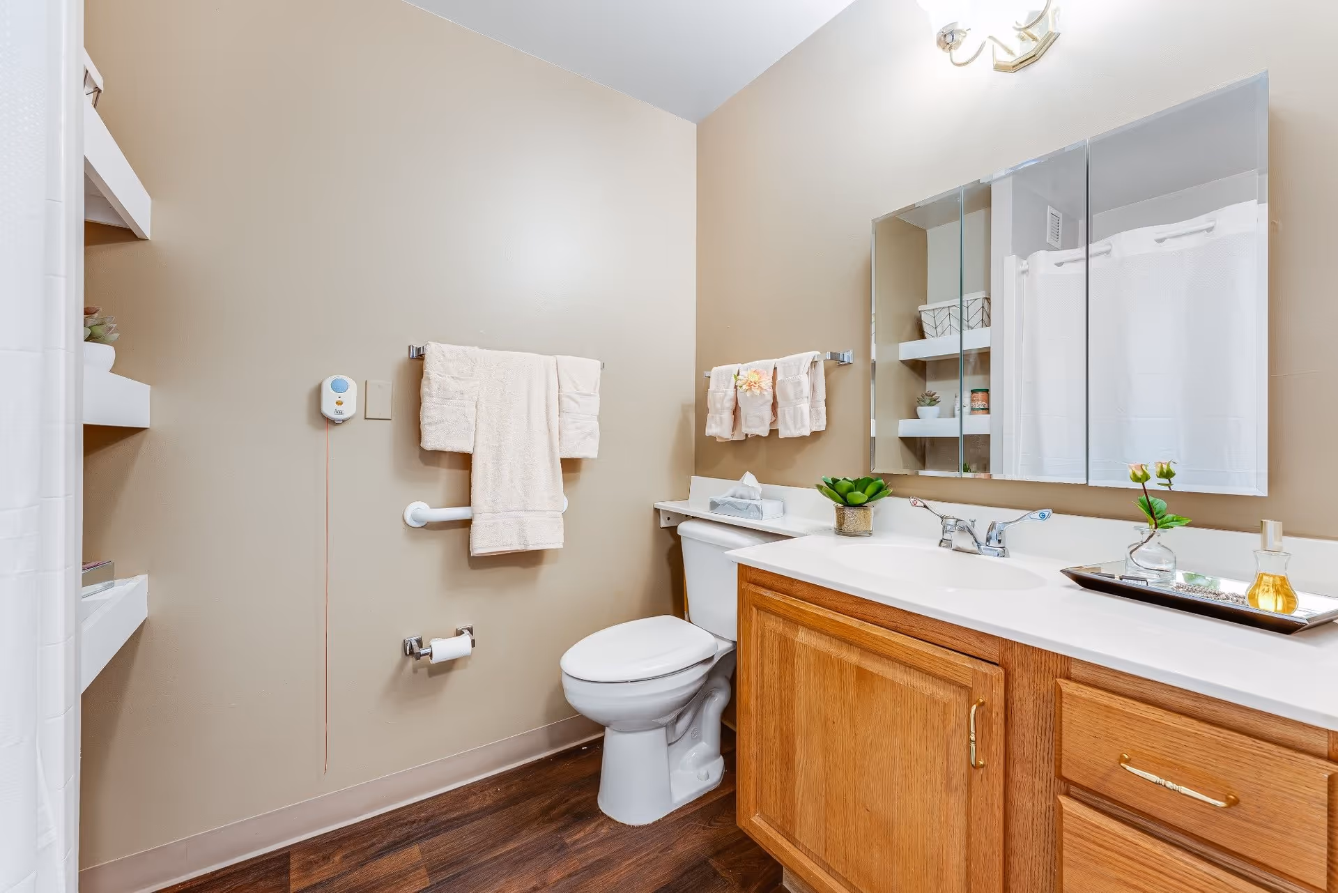 A clean bathroom with beige walls and wood flooring. The room features a white toilet, a wooden vanity with a white countertop and a sink, a large mirror above the sink, and a towel rack with beige towels. There are shelves with decorative items and a white shower curtain visible on the left side.