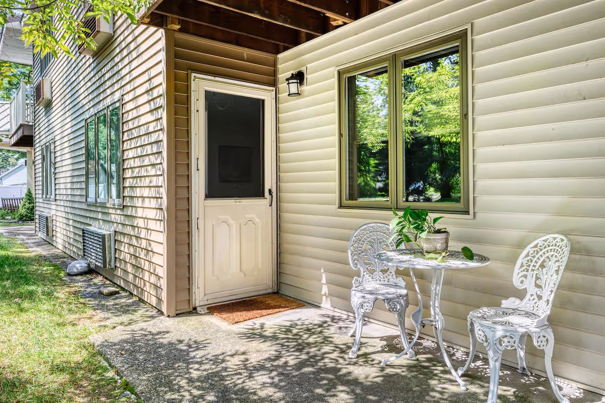 Small outdoor patio beside a beige-sided building with a white screen door, window, and a white metal bistro table with two chairs.