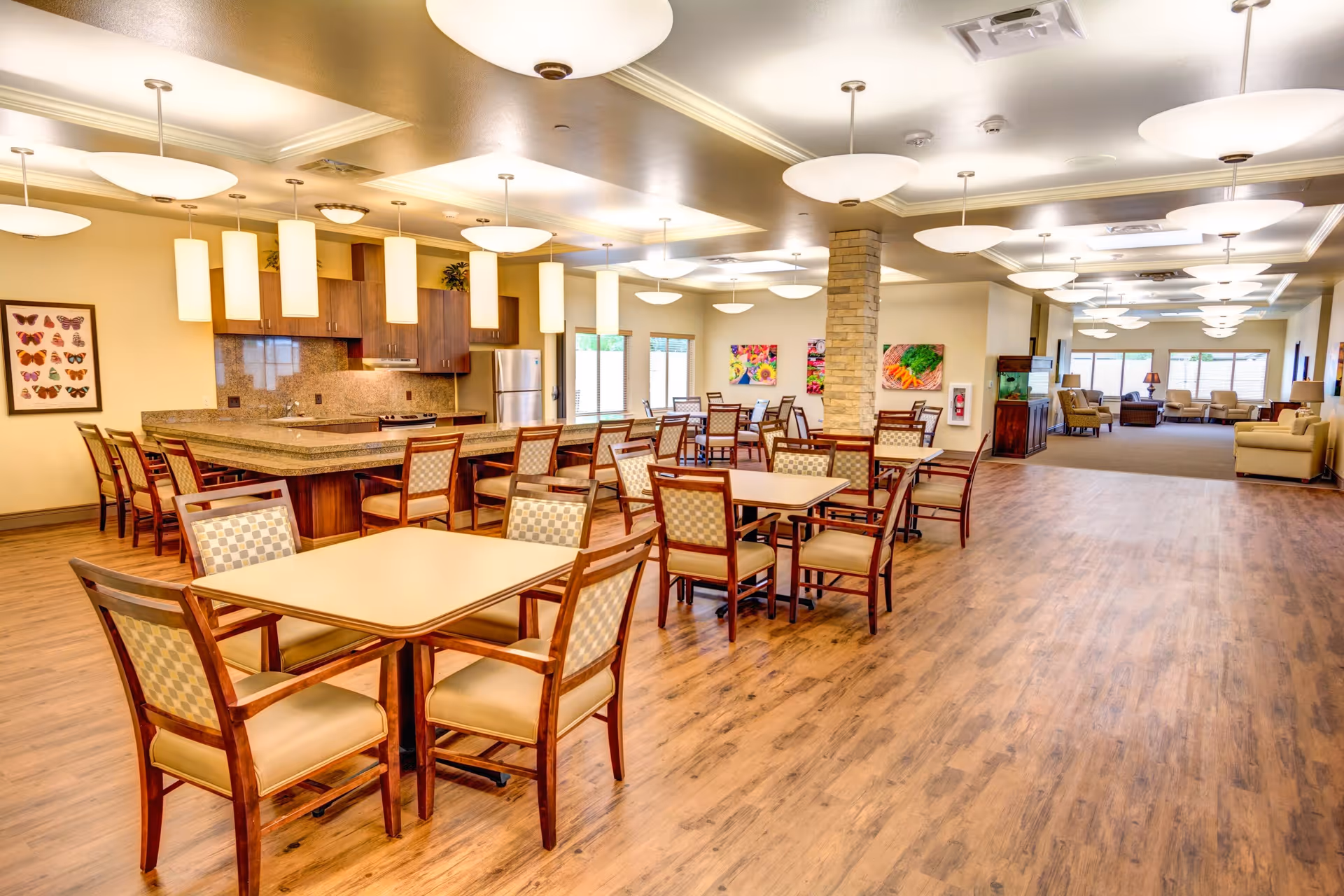 A spacious dining area in a senior living facility featuring multiple wooden tables with cushioned chairs arranged neatly. The room has a kitchen area with a granite countertop island, wooden cabinets, and stainless steel appliances. The floor is wood, and the ceiling has modern light fixtures. In the background, there is a lounge area with sofas and large windows allowing natural light to enter.