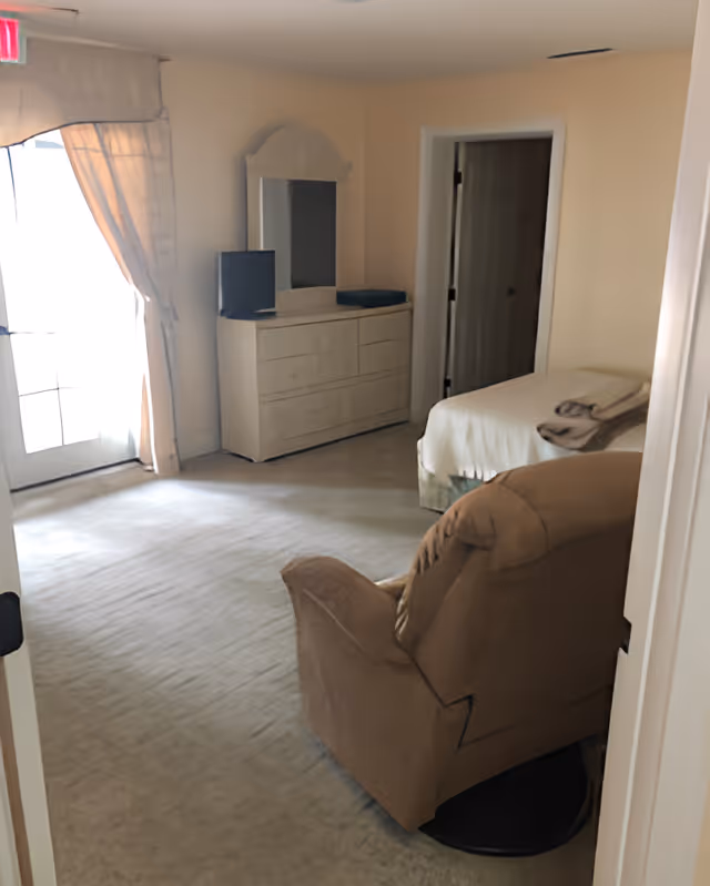 A bedroom with a beige armchair in the foreground, a bed with folded towels on it, a dresser with a mirror, and a door leading to another room. There is a window with curtains letting in natural light.