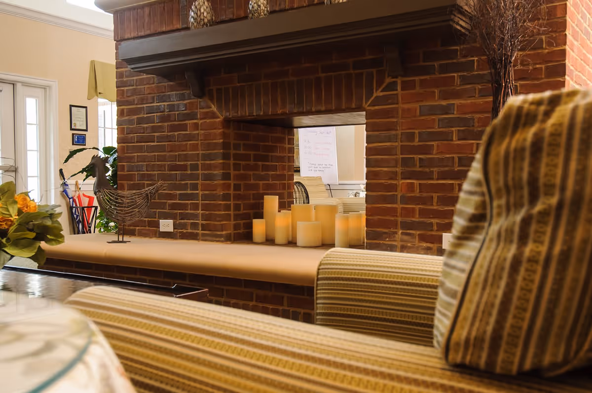 Cozy interior view of a senior living facility featuring a brick fireplace with multiple pillar candles on the hearth. In the foreground, there is a striped upholstered chair. The background shows a door with glass panels, some plants, and a decorative metal rooster sculpture on the hearth.