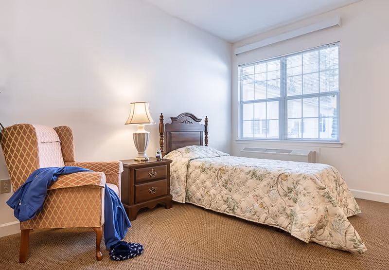 A simple furnished bedroom with a twin bed, floral bedspread, wooden nightstand and lamp, an upholstered armchair, and a large window.