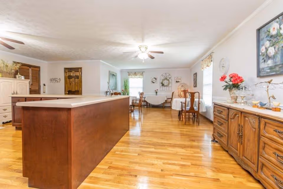 A spacious room with polished hardwood floors featuring a large wooden kitchen island in the foreground. In the background, there are several small dining tables with chairs, decorated with tablecloths and floral arrangements. The room has white walls, windows with floral curtains, ceiling fans with lights, and various decorative items including paintings and plants.