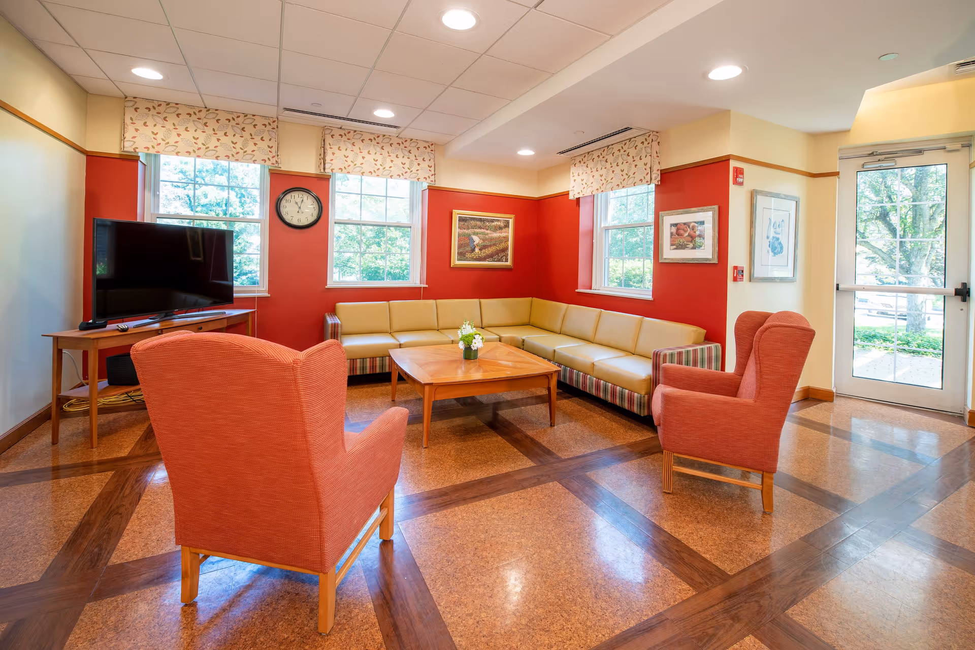 A cozy living room area with a large L-shaped beige sofa, two orange armchairs, a wooden coffee table with a small flower arrangement, a flat-screen TV on a wooden stand, three windows with patterned valances, and framed artwork on red and cream walls. The room has a polished floor with a wood inlay pattern and a glass door leading outside.