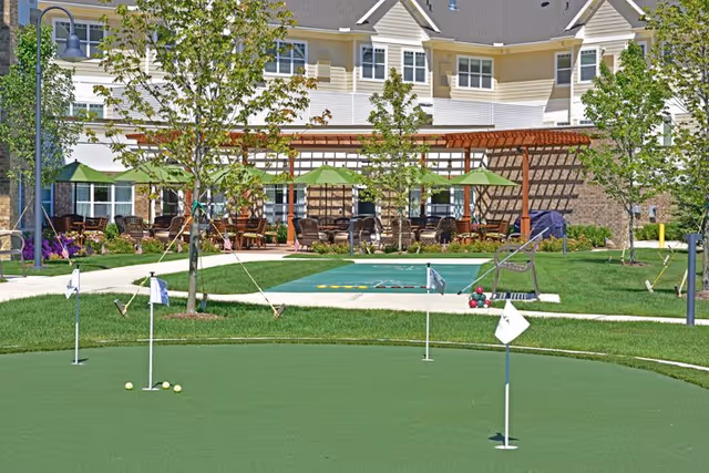 Outdoor recreational area at Rose Senior Living Avon featuring a putting green with golf flags and balls, a shuffleboard court, patio seating with tables and green umbrellas, surrounded by trees and a multi-story building in the background.