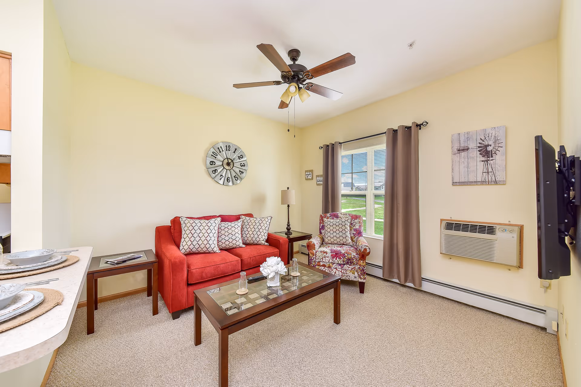 A cozy living room with a red sofa adorned with patterned cushions, a floral armchair, a glass-top coffee table with decorative items, a wall clock, a ceiling fan with lights, a window with brown curtains, and a wall-mounted air conditioning unit.