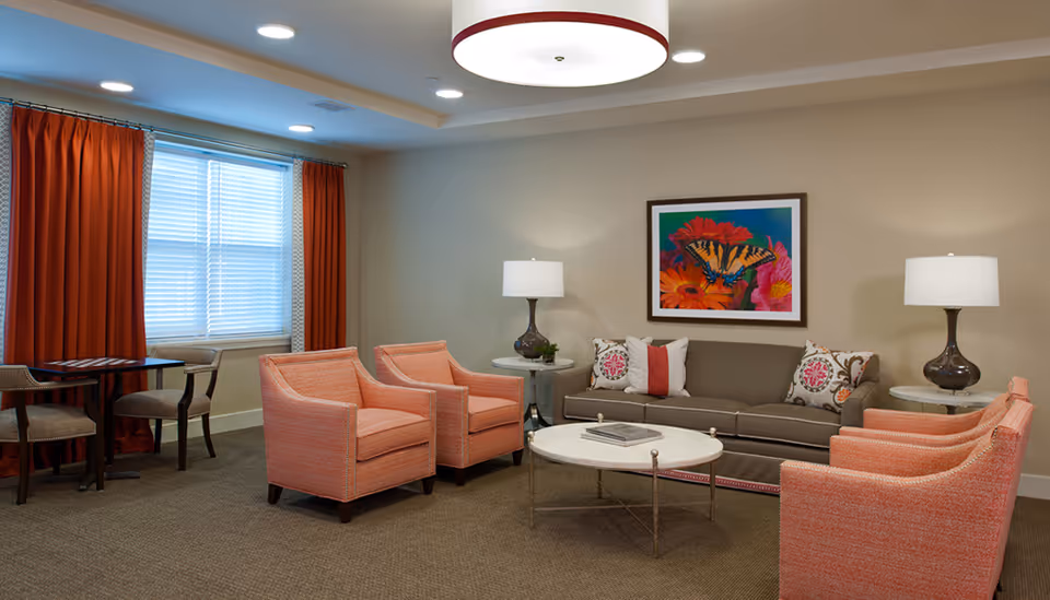 A cozy living room with a gray sofa adorned with decorative pillows, four coral-colored armchairs, two round white side tables with lamps, a round white coffee table with books, a window with orange curtains, and a colorful butterfly artwork on the wall.