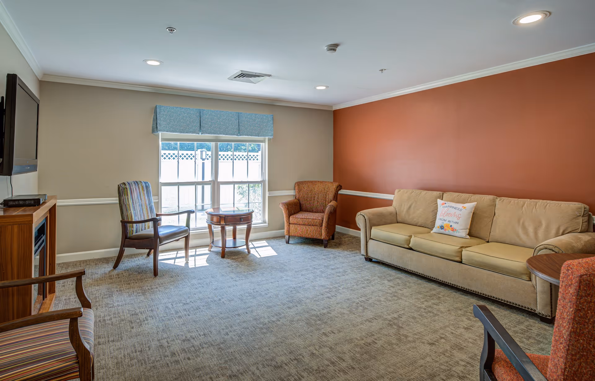 A cozy living room with a beige couch, an orange armchair, two wooden chairs with striped cushions, a small round wooden table, a wall-mounted TV, and a large window with a blue valance allowing natural light to fill the room. The walls are painted beige and one accent wall is painted burnt orange.