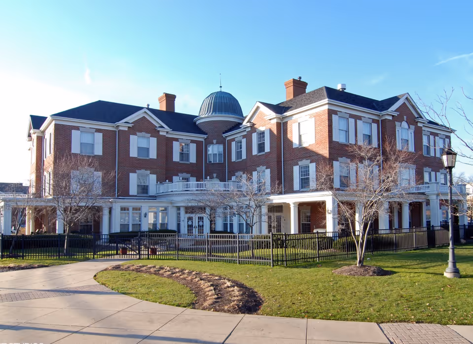 Exterior view of a large, three-story brick building with white window shutters and a distinctive dome-shaped roof feature. The building is surrounded by a black metal fence, leafless trees, a lamppost, and a well-maintained lawn with a curved sidewalk leading up to the entrance.