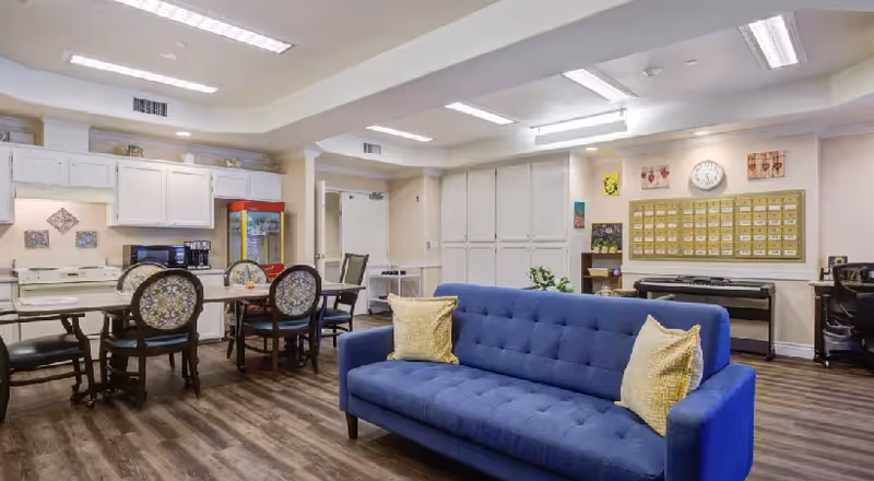 Interior view of a senior living facility common area with a blue sofa with two yellow pillows in the foreground, a dining table with chairs, white cabinets, a popcorn machine, and a wall with a clock and mailboxes in the background.