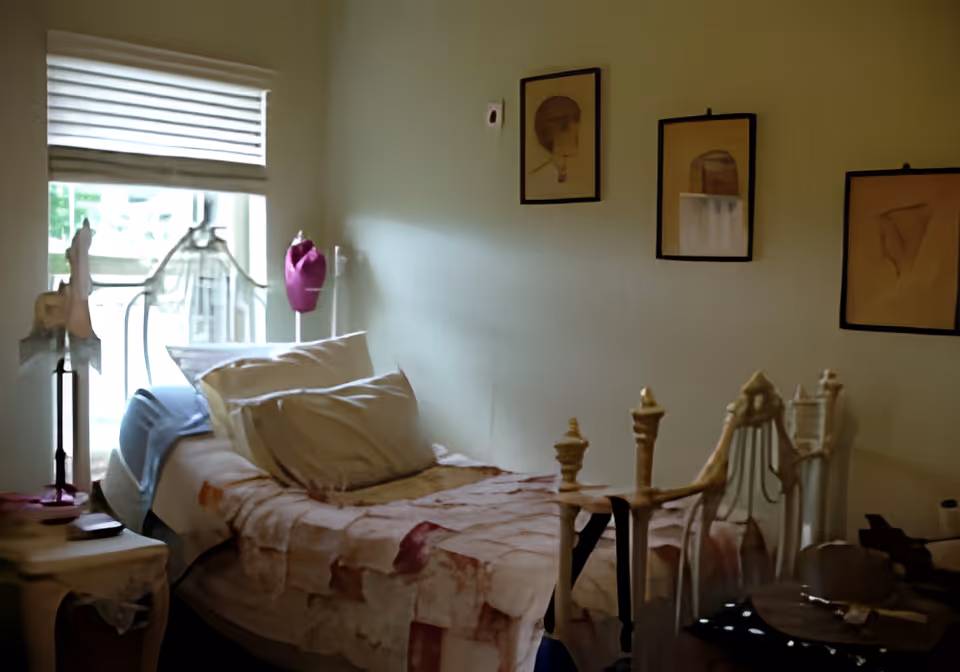 A small bedroom with an ornate metal single bed dressed with pillows and a patterned bedspread, a window with blinds, framed artwork on the wall, and bedside tables.