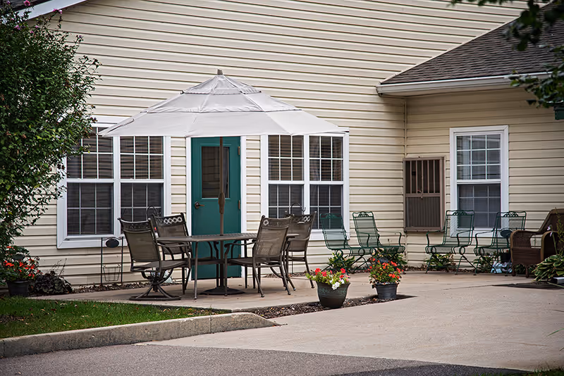 Patio with a round table, umbrella, multiple chairs and potted flowers in front of a beige-sided building.